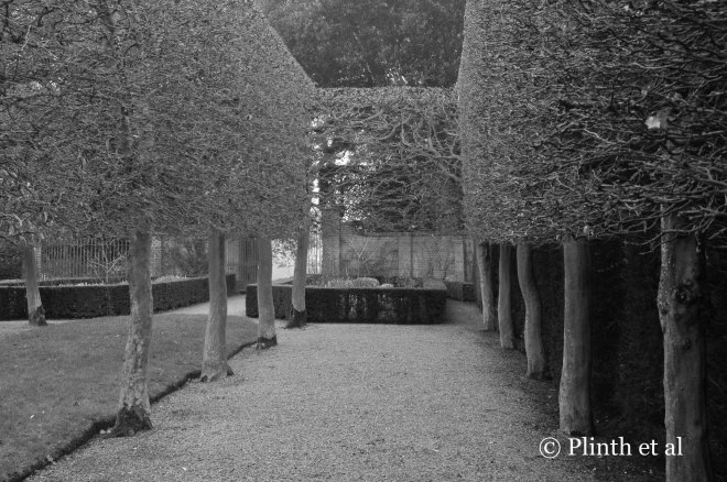 Denuded of their leaves, the pleached hornbeams become wiry edifices that play off texturally the solid boxwood and yew hedges, and the grass panel, walls, and gravel paths are tonally different from the clipped plants.